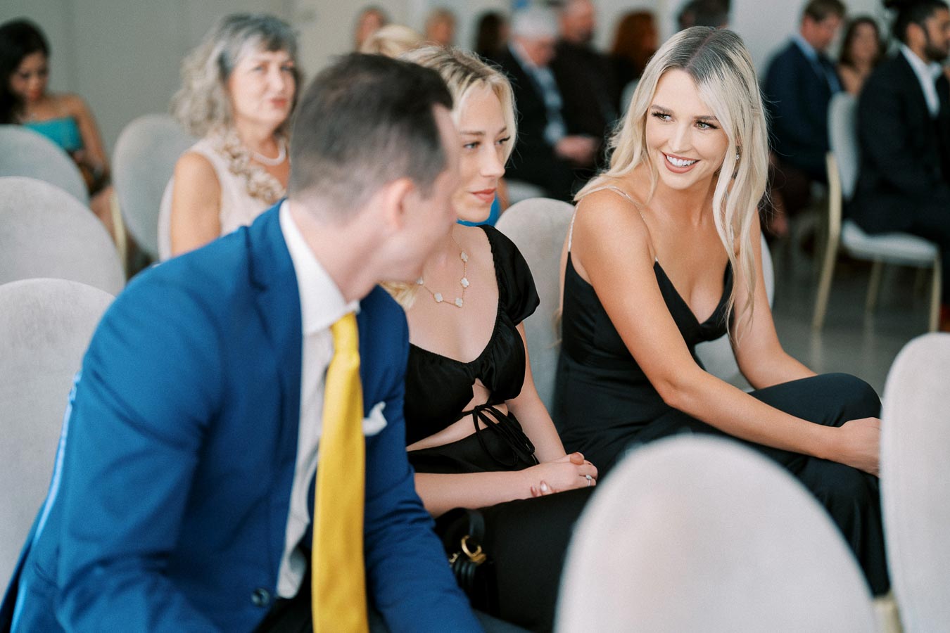 A group of elegantly dressed people sitting and conversing at a formal event, with a woman in a black dress smiling warmly and a man in a blue suit with a yellow tie attentively listening.