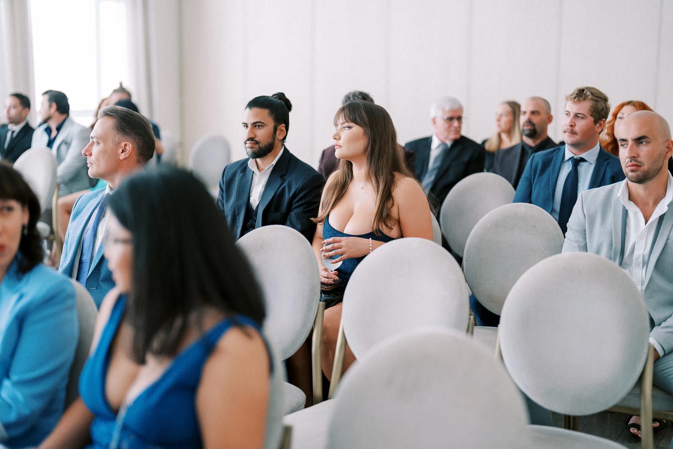 Alt text: Audience seated in a formal event, with people dressed in suits and elegant attire, listening attentively in a bright, modern venue.