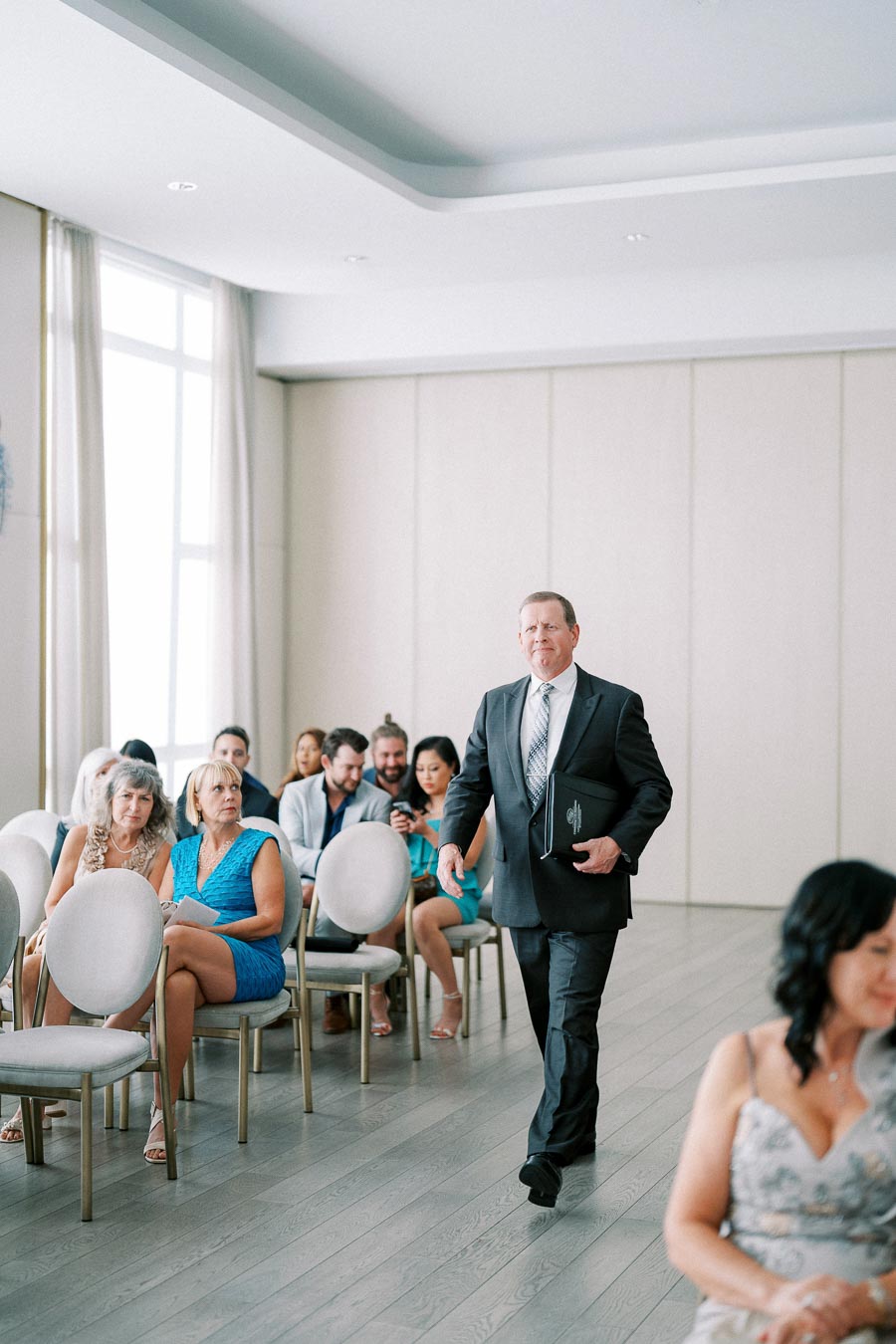A man in a suit walks down an aisle in a modern, sunlit conference room, surrounded by seated guests.