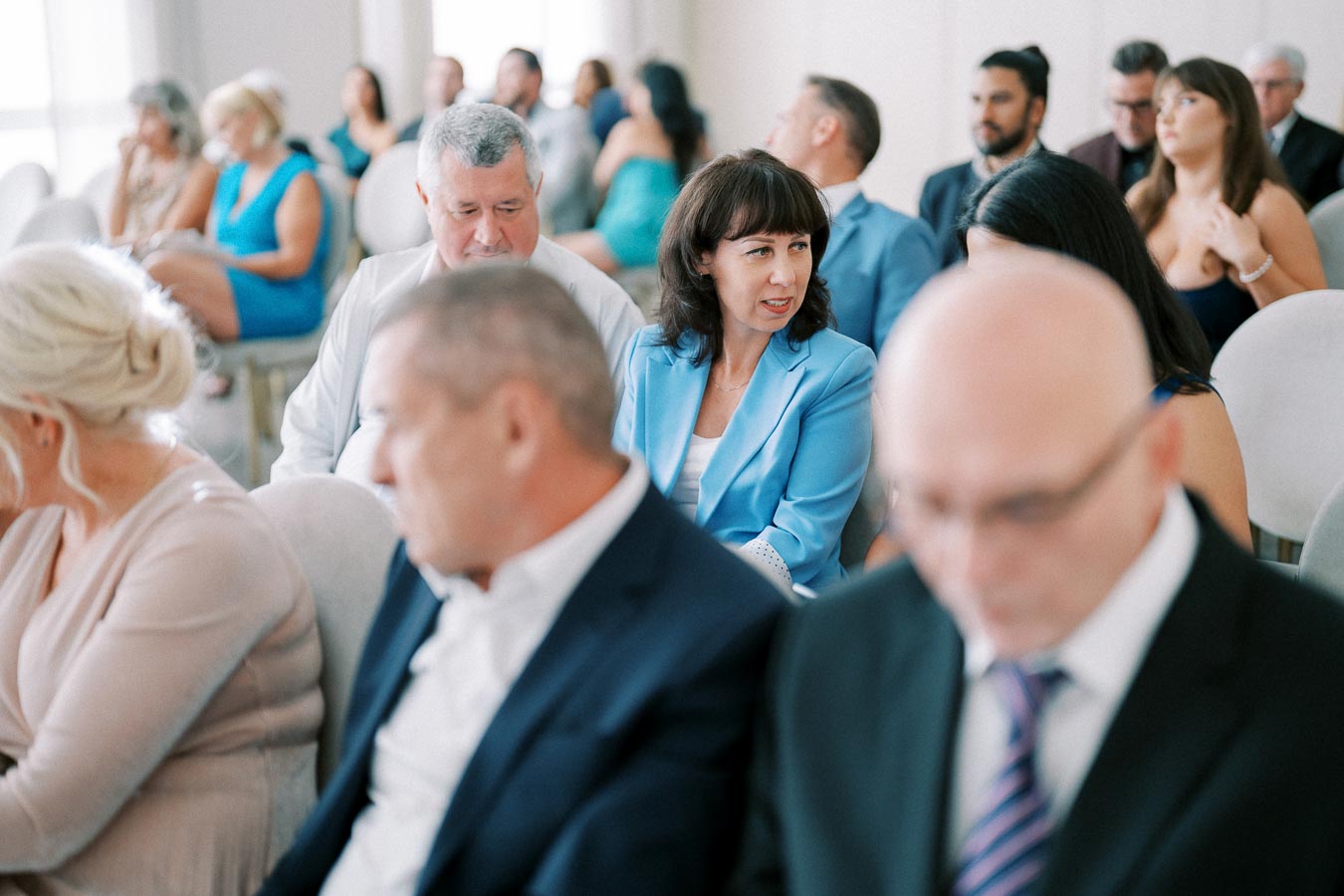 A diverse group of people in formal attire seated indoors at a gathering, with a woman in a blue blazer engaging in conversation, suggesting an event or conference setting.