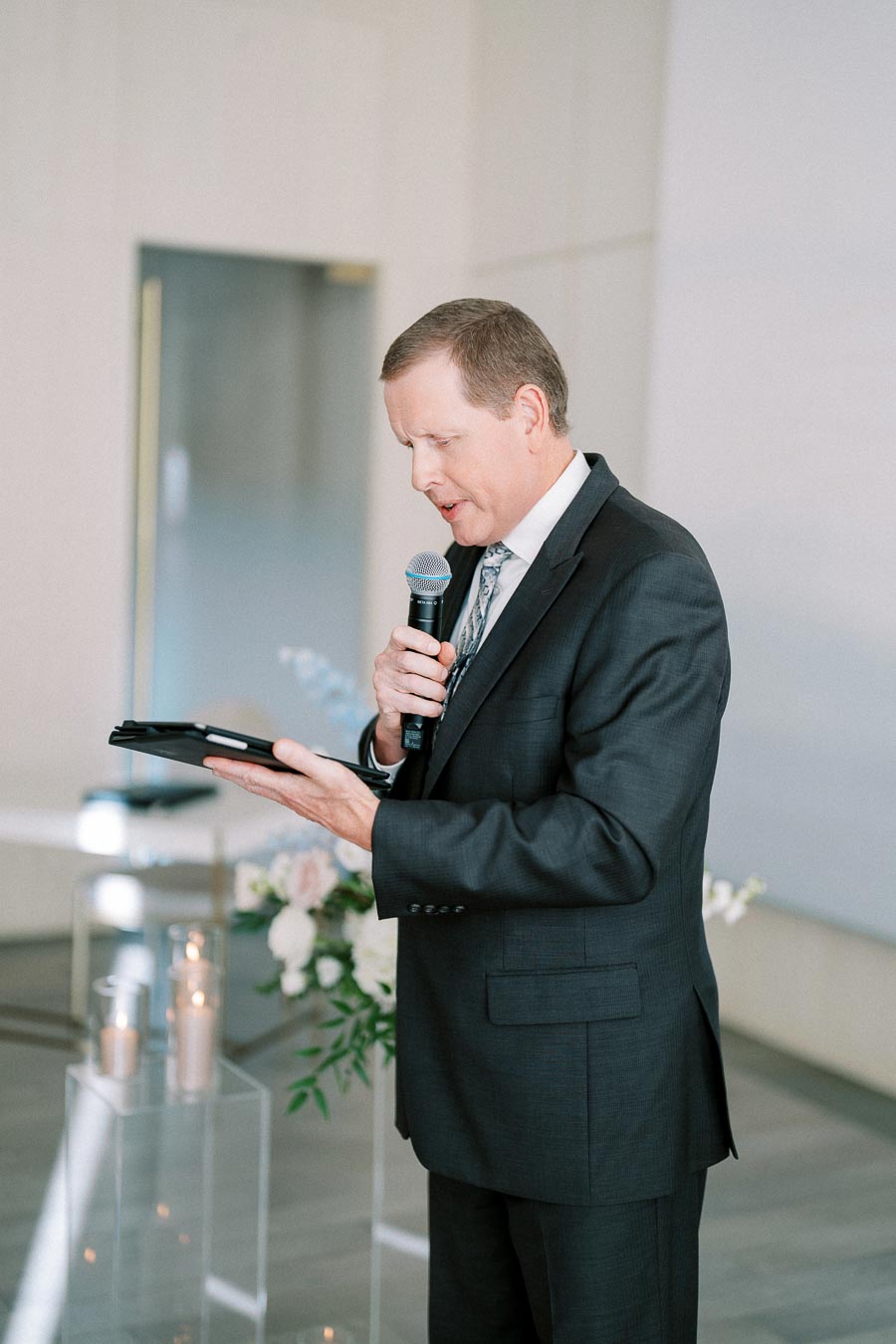 A man in a suit giving a speech into a microphone while holding a tablet, with a floral arrangement and candles in the background at an indoor event.