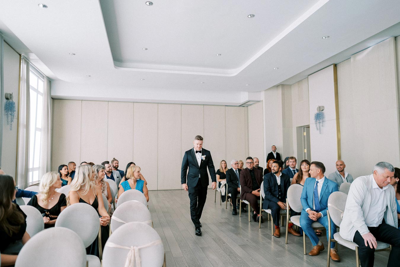 A man in a black suit and bow tie walking down the aisle of a modern wedding ceremony venue, with seated guests watching attentively, in a bright room with large windows.