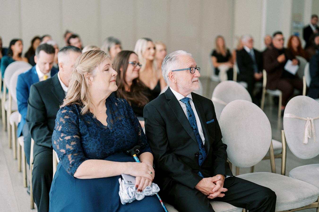 Elderly couple sitting in an elegant indoor wedding ceremony, surrounded by guests dressed in formal attire.