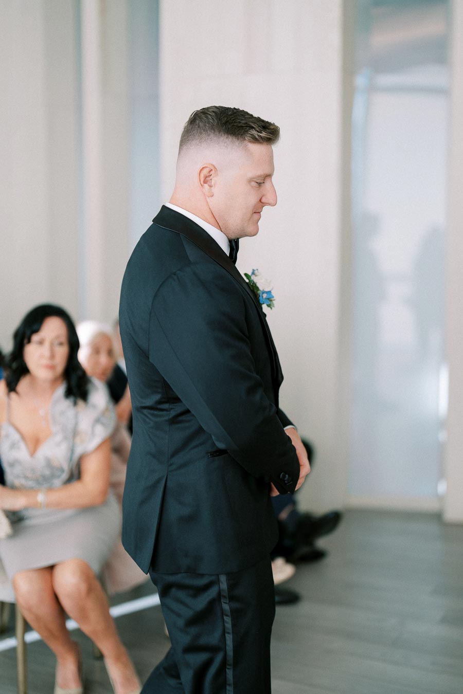 Man in a black suit standing indoors, with seated guests in the background.
