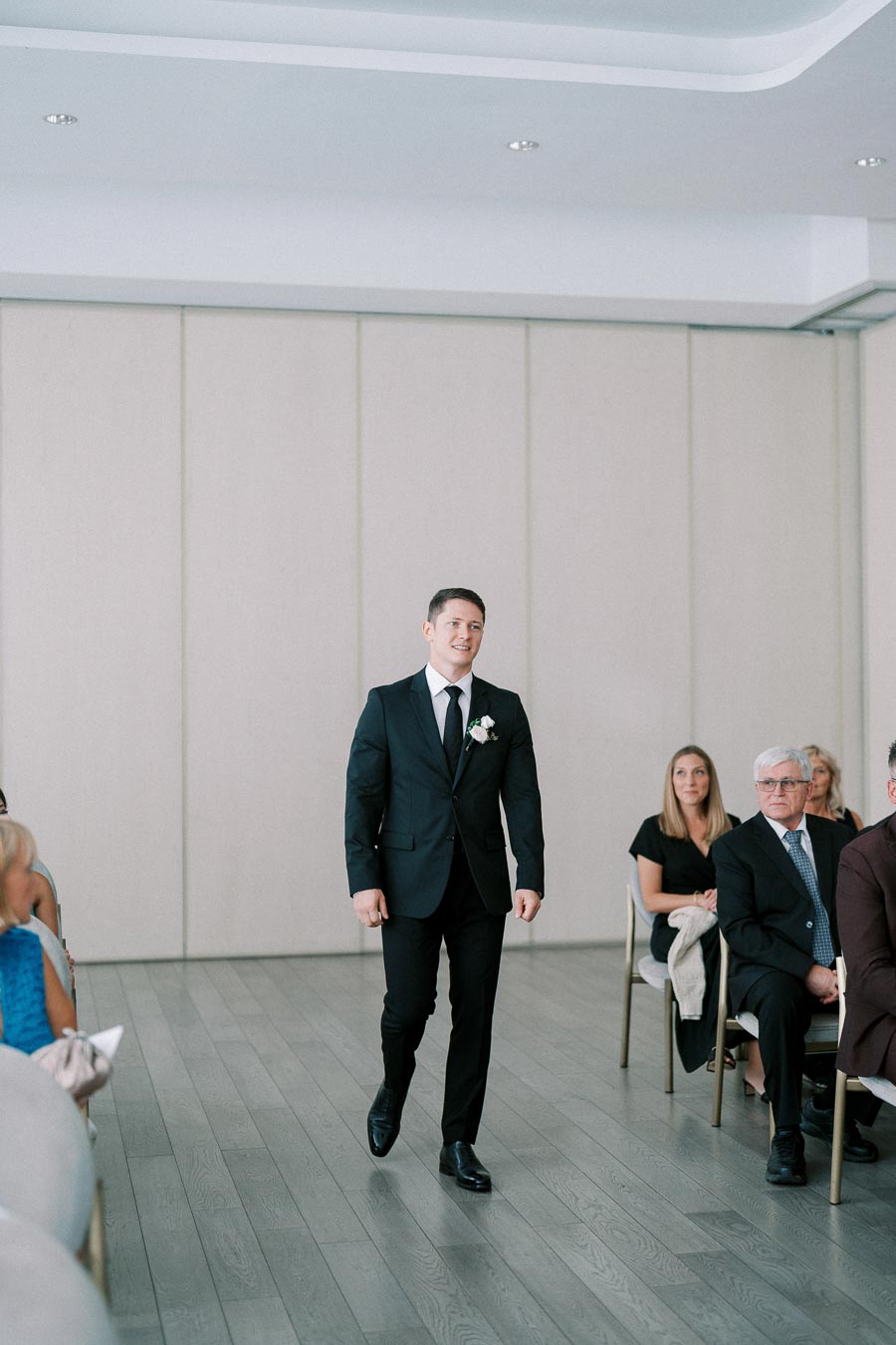 A man in a formal black suit and tie walking down an aisle during a wedding ceremony, with guests seated on chairs watching.