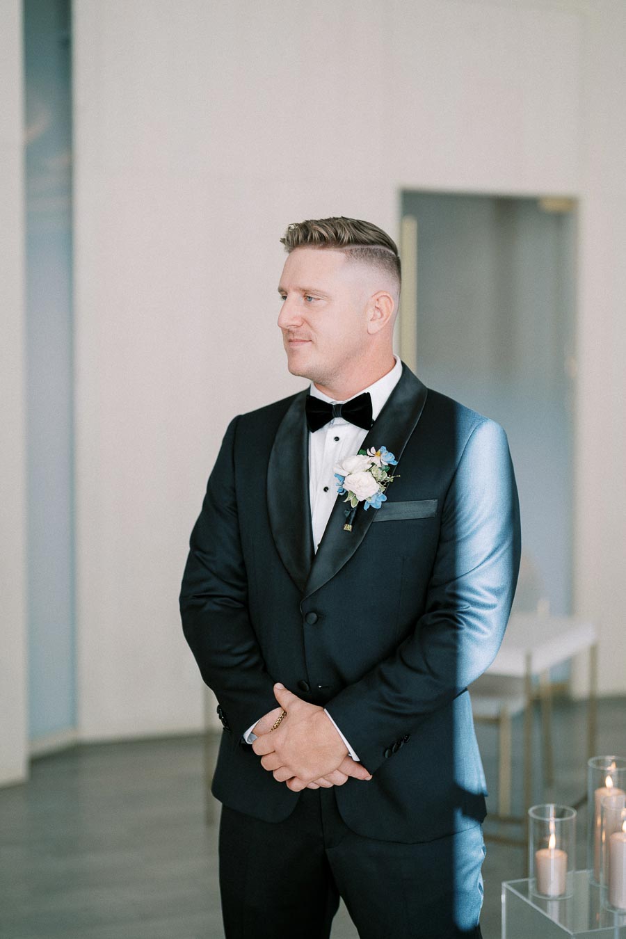 A groom in a black tuxedo with a white shirt and bow tie, adorned with a boutonniere, standing indoors with a serene expression amidst soft, ambient lighting.