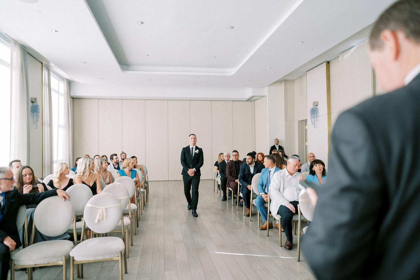 A man in a suit walking down the aisle of a bright, modern wedding venue, surrounded by seated guests dressed in formal attire, with large windows allowing natural light to fill the room.