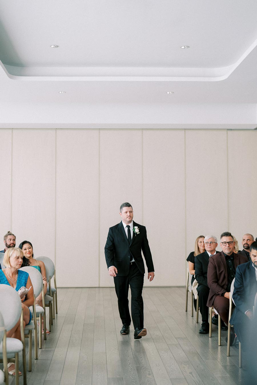 A man in a black suit and tie walks down the aisle of a modern wedding venue, with guests seated on either side, creating a formal and elegant atmosphere.