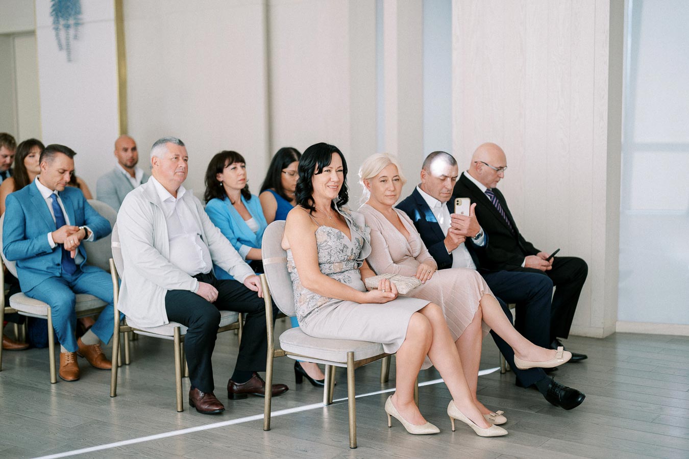 A group of people sitting in a formal event, including women in elegant dresses and men in suits, some using smartphones, in a well-lit room with chairs arranged in rows.