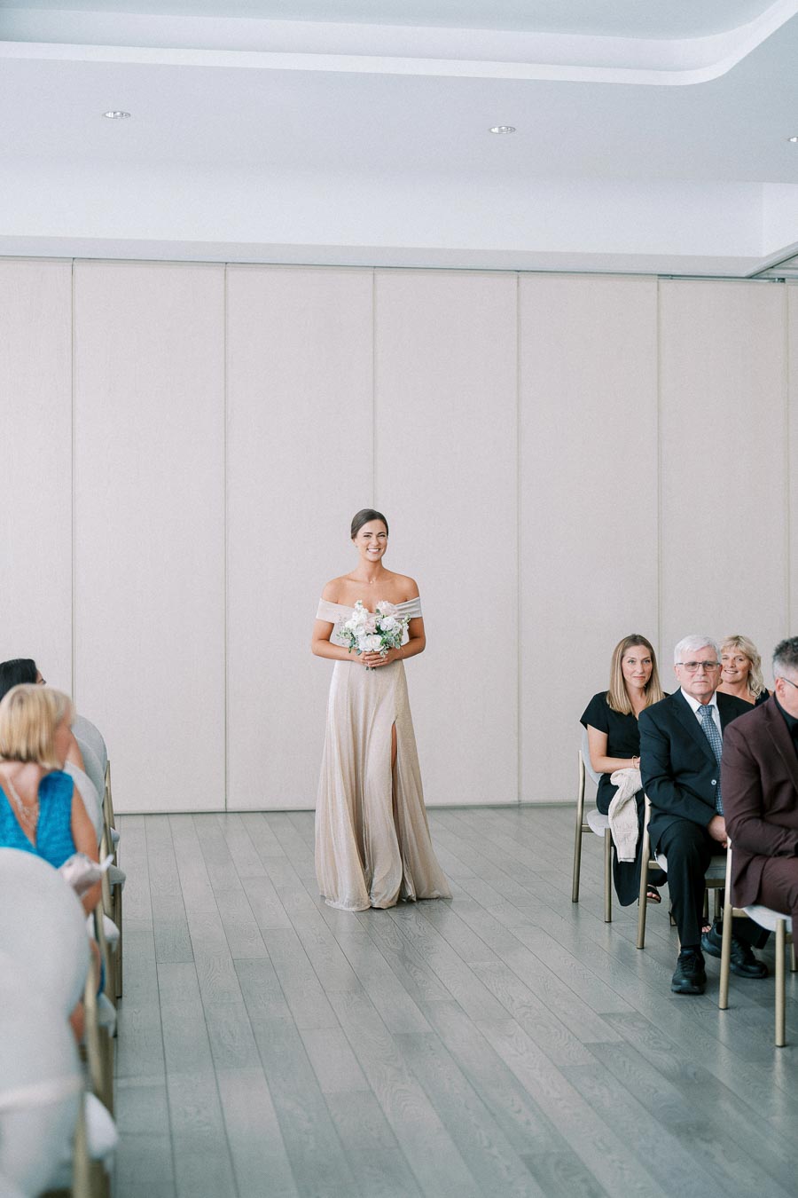 Bridesmaid in a flowing beige gown walking down the aisle holding a bouquet, surrounded by seated wedding guests in a brightly lit venue.