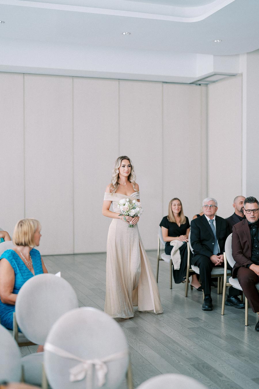 A bridesmaid in an elegant off-shoulder gown walks down the aisle during a wedding ceremony, holding a bouquet of white flowers in a modern indoor venue, with seated guests in the background.
