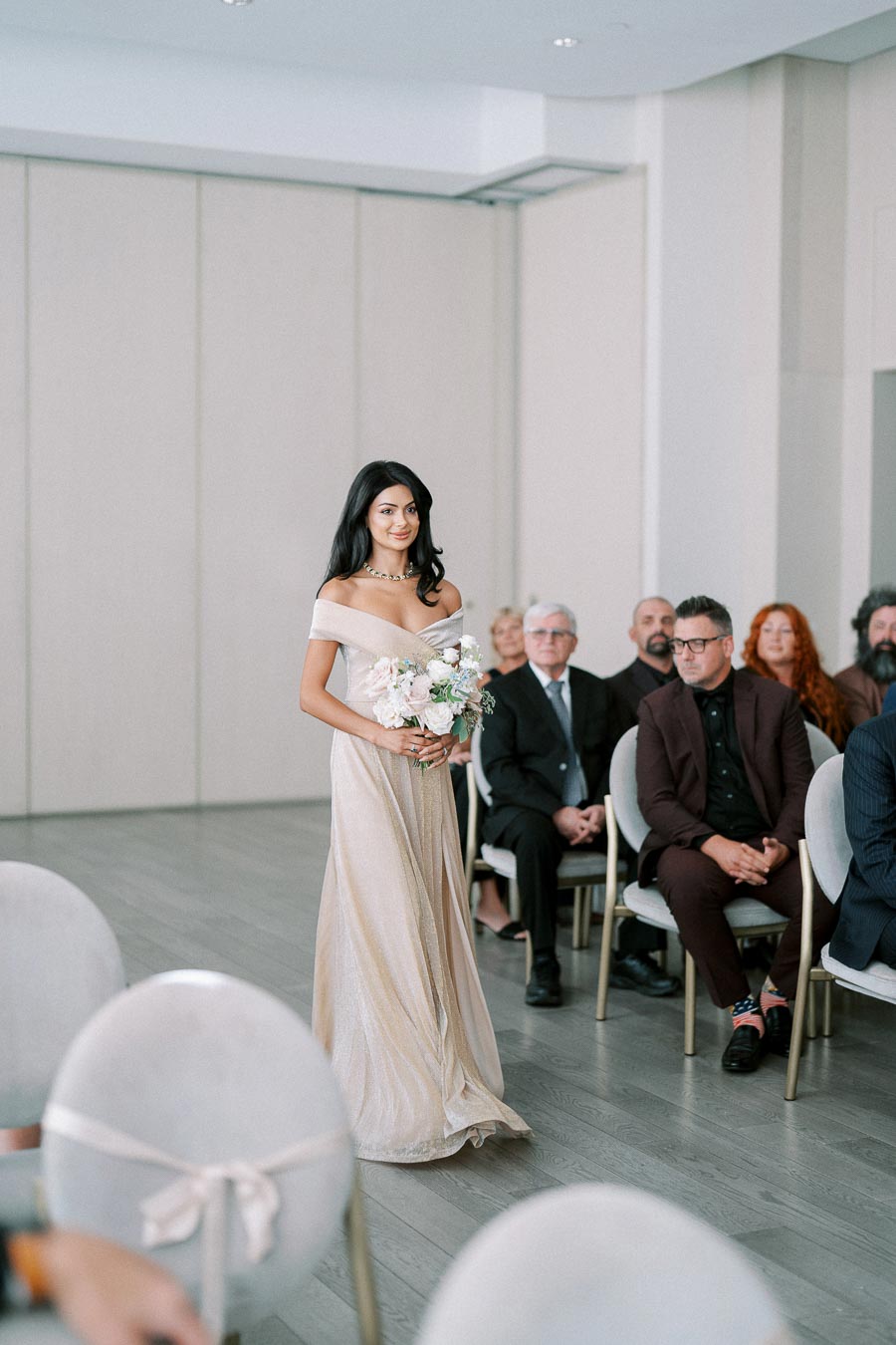 Bridesmaid walking down the aisle in an elegant off-shoulder gown, holding a pastel floral bouquet, with seated guests in the background.