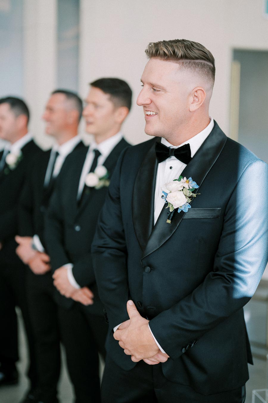 A groom and his groomsmen in matching black tuxedos with boutonnières, standing and smiling during a wedding ceremony.