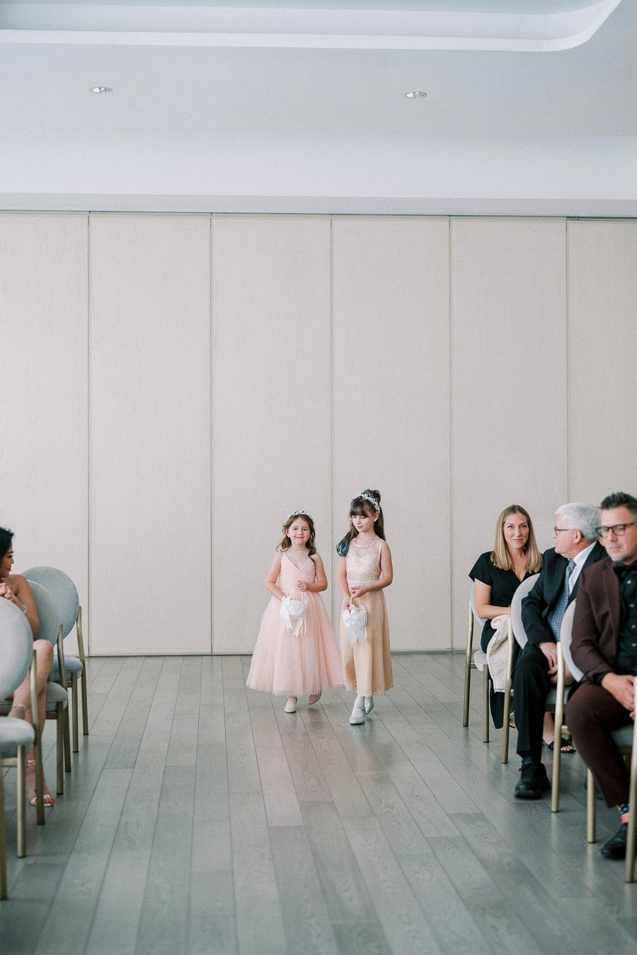 Two young flower girls in elegant peach dresses walking down an aisle during a wedding ceremony, carrying white baskets, with seated guests observing in a modern venue.