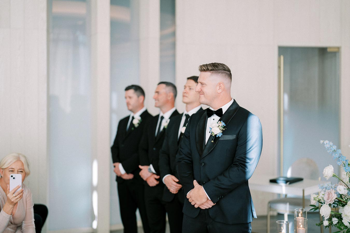 Groom and groomsmen in formal suits stand together at an indoor wedding ceremony, with white flower boutonnieres and a woman taking a photo in the foreground.