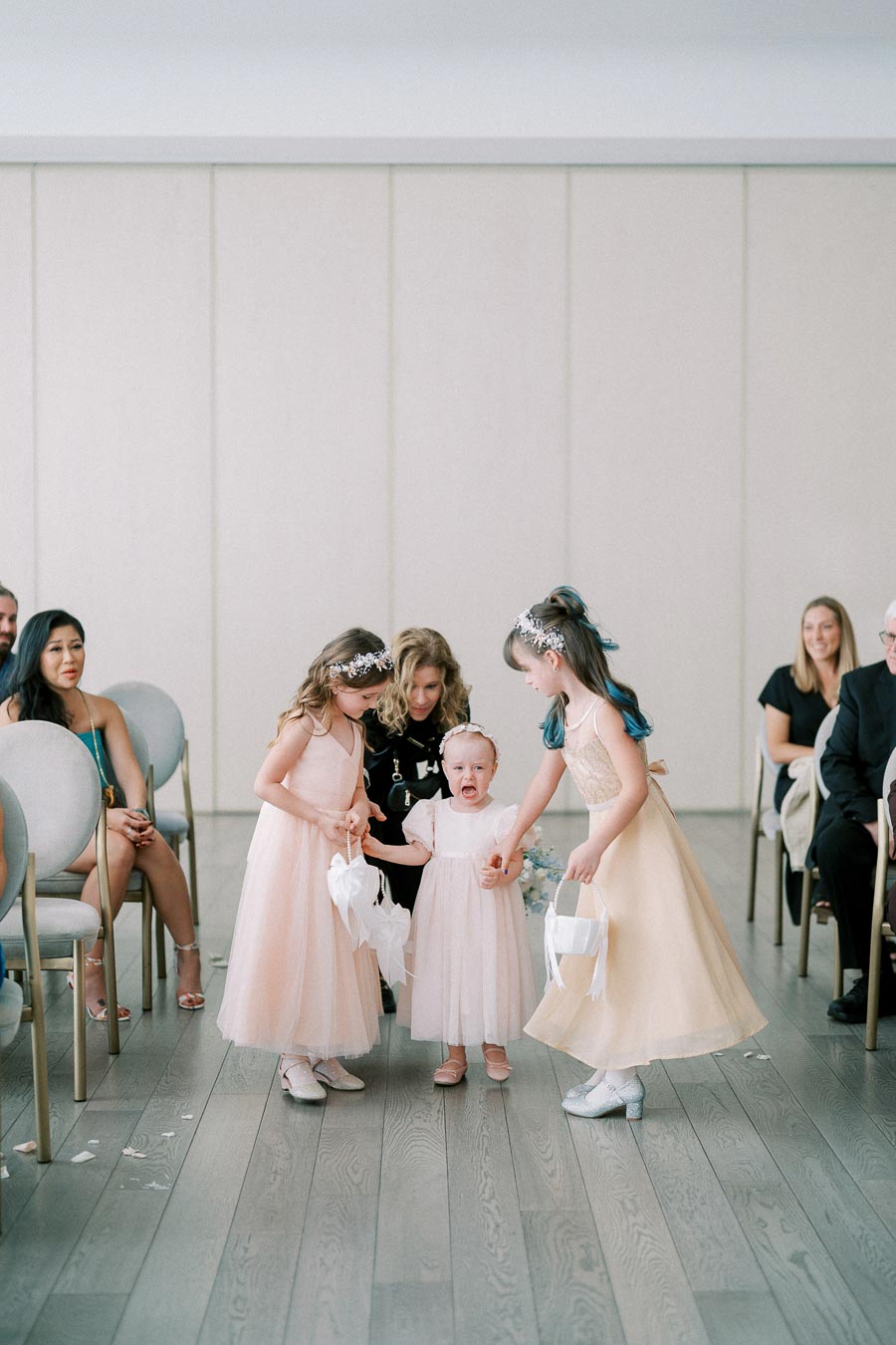 Cute flower girls in peach dresses participating in a wedding ceremony, with one child crying, surrounded by seated guests on a wooden floor.