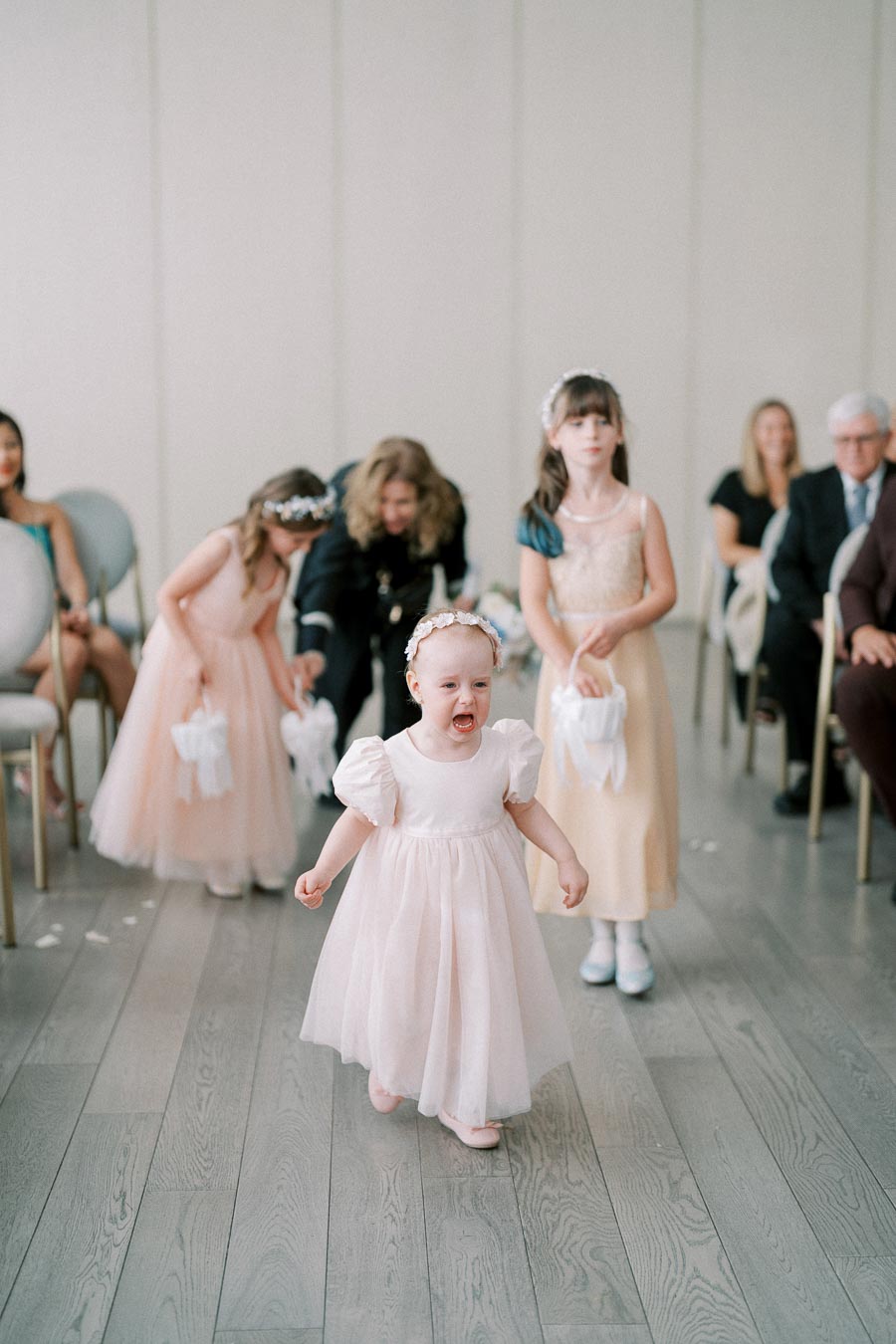 Cute toddler in a pink dress crying during a wedding ceremony with flower girls and guests in the background.