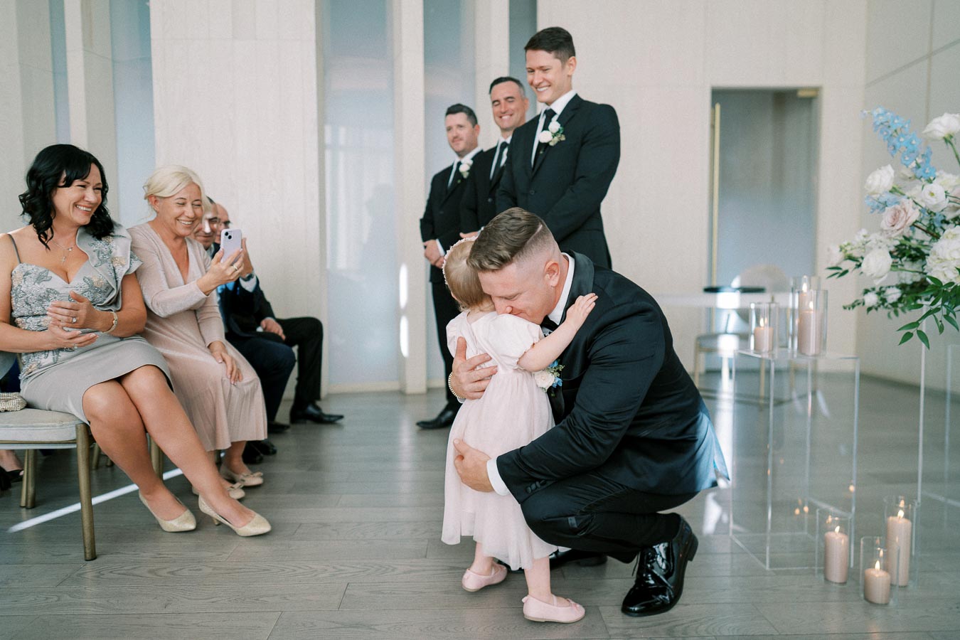 A groom in a black suit lovingly embraces a young girl in a pink dress during a wedding ceremony, surrounded by smiling guests in a bright, elegantly decorated venue.