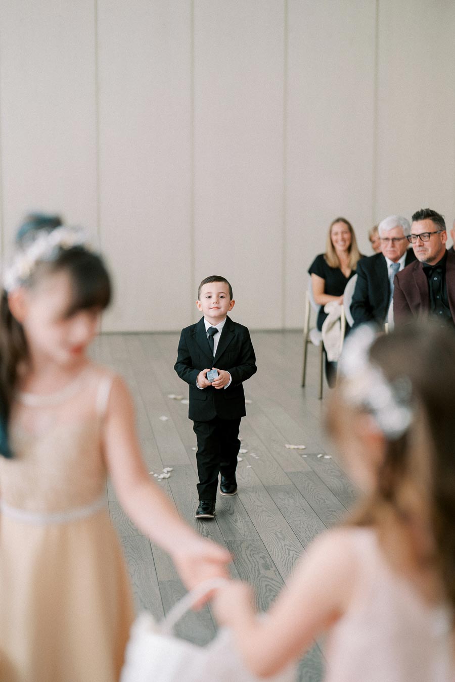 A young boy in a black suit walks down the aisle at a wedding ceremony, holding a ring box. Blurred flower girls in dresses are in the foreground, with seated guests smiling in the background.