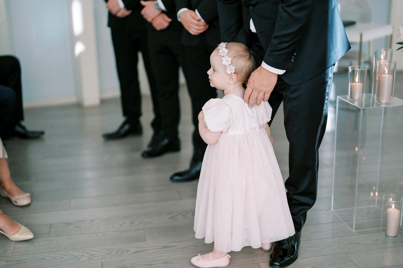 A young child in a white dress and headband stands next to an adult in formal attire, holding their hand. The scene includes other people dressed in suits and elegant shoes, suggesting a formal gathering or event. Lit candles in glass holders are visible, enhancing the serene ambiance.