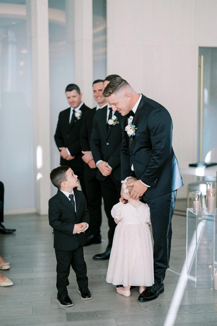 A groom in a navy suit interacts warmly with a young ringbearer and flower girl during a modern wedding ceremony indoors.