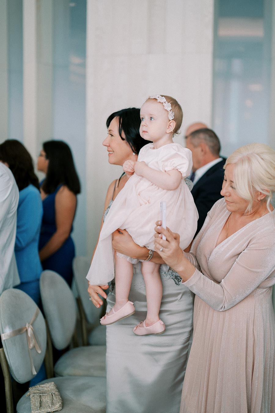A woman holding a baby girl in a pink dress and headband at a formal event, surrounded by other attendees in elegant attire.