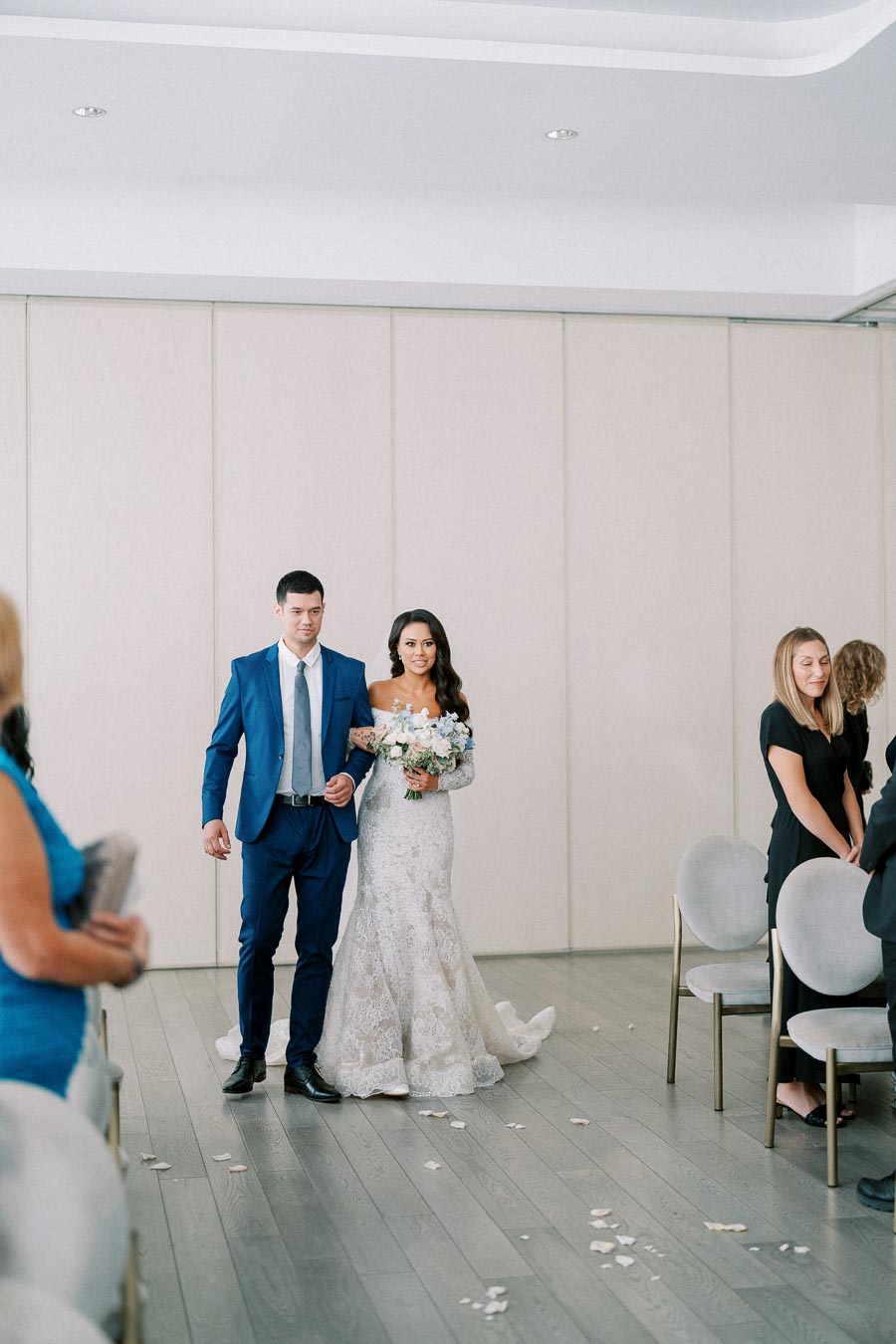 Elegant bride in lace gown holding a bouquet, walking down the aisle escorted by a man in a blue suit during a wedding ceremony.