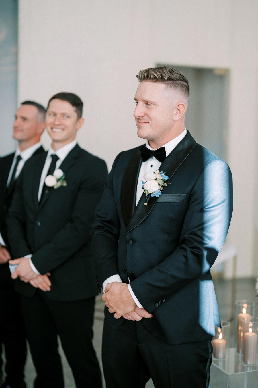 Groom in a navy tuxedo with a boutonniere stands confidently at a wedding ceremony, accompanied by groomsmen in black suits smiling in the background.