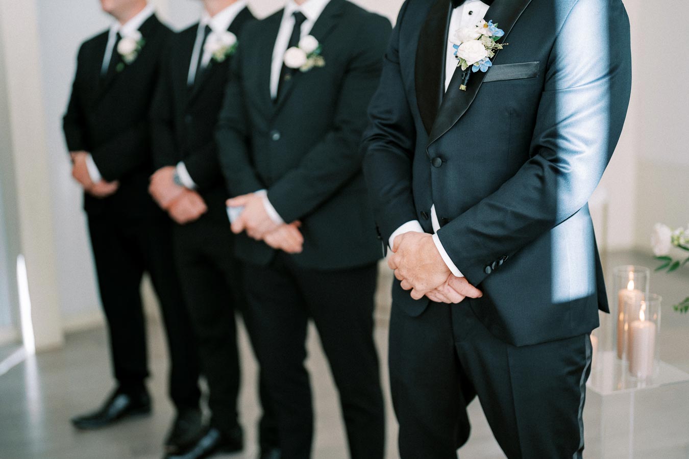 A group of groomsmen in elegant black suits and white boutonnieres, standing in a row at a wedding ceremony.