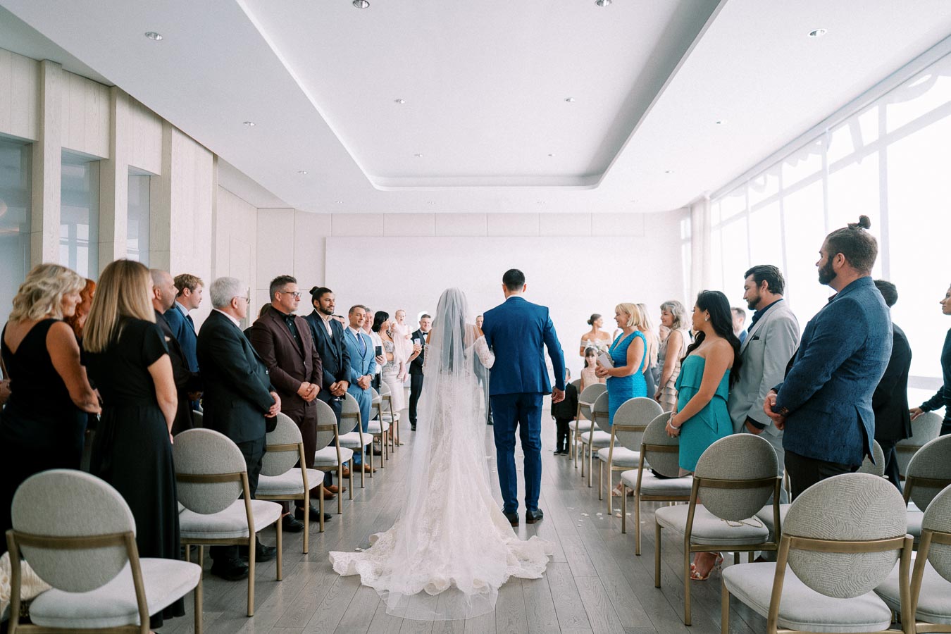 A bride and groom walk down the aisle in a modern, bright wedding ceremony with guests standing on either side. The venue features high ceilings and a minimalist design, creating an elegant atmosphere.