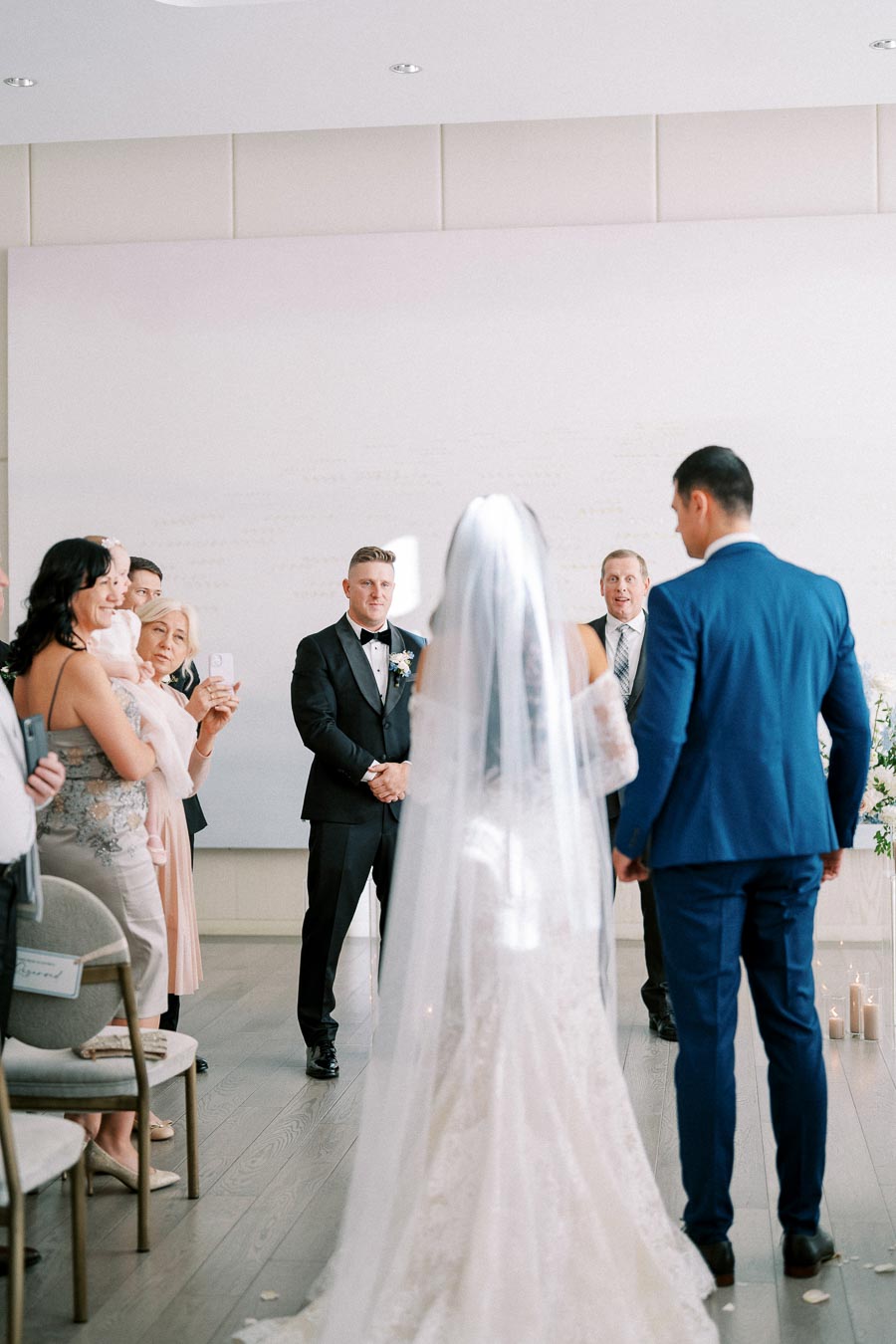 Wedding ceremony with bride walking down the aisle towards groom in a black tuxedo, surrounded by smiling guests capturing the moment on their phones. Elegant indoor venue with soft lighting and floral decorations.