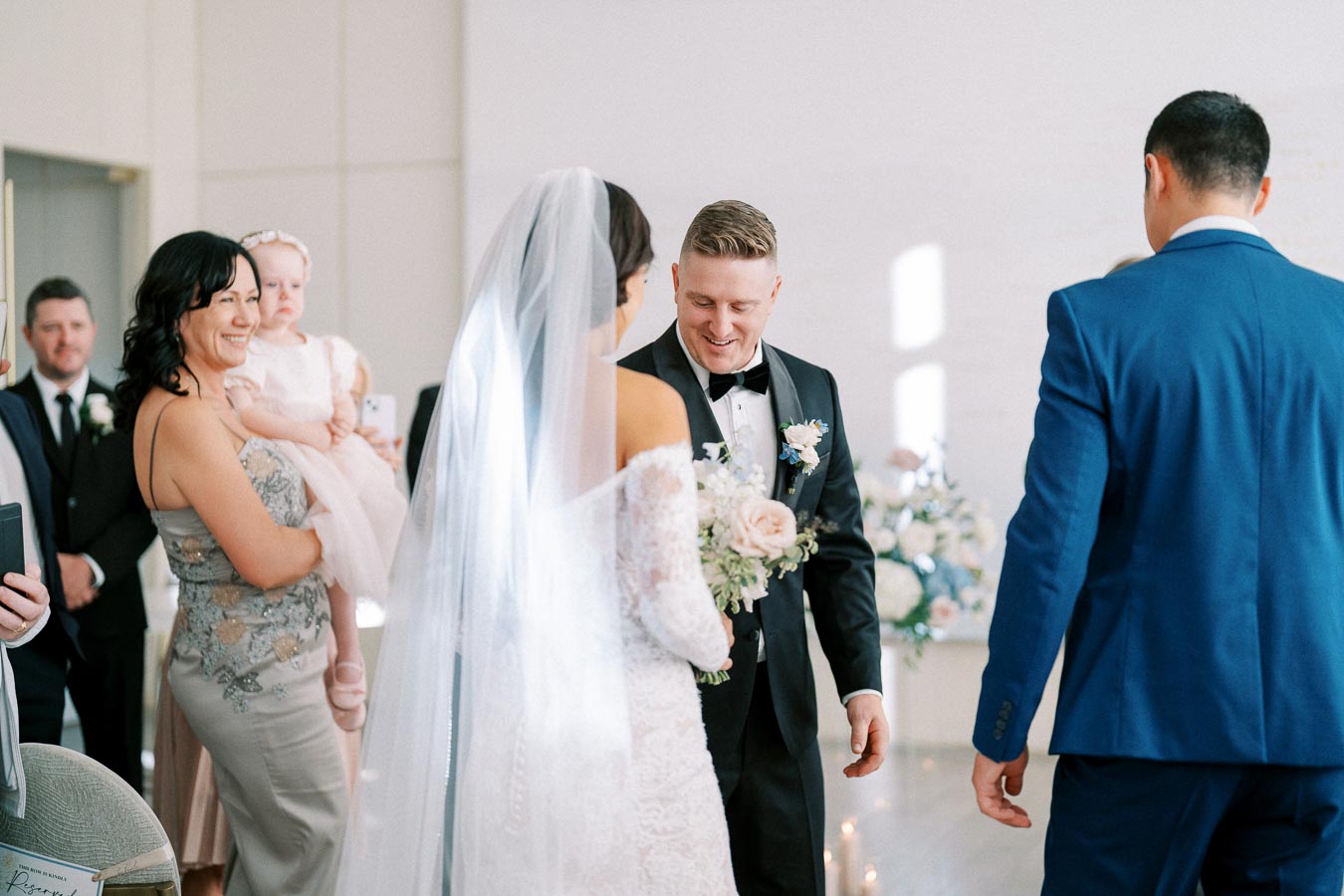A bride and groom exchanging smiles during their wedding ceremony, surrounded by elegantly dressed guests, with a mother holding a baby in the background.