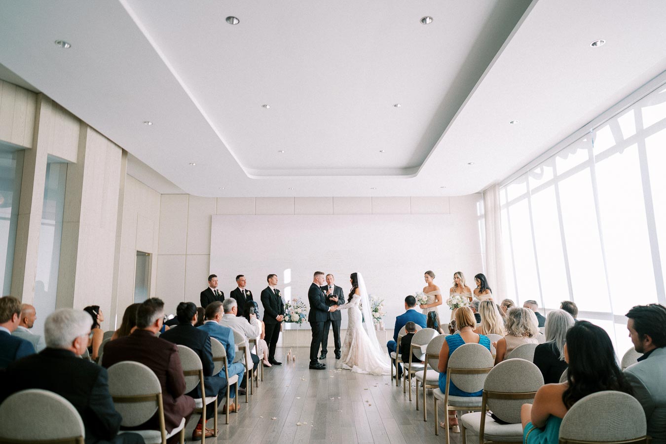 A wedding ceremony takes place in a modern, light-filled room. The bride and groom stand at the altar surrounded by bridesmaids and groomsmen, as guests seated in rows watch the event.