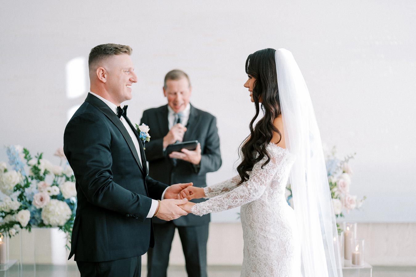 Bride and groom smiling and holding hands during a wedding ceremony, with an officiant reading from a book in a bright, floral-decorated venue.