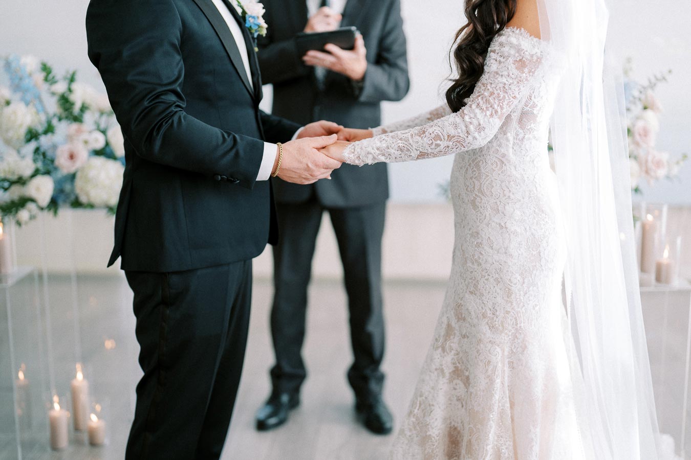 Elegant wedding ceremony with a bride in a lace gown and groom in a black suit holding hands, surrounded by floral arrangements and candles.