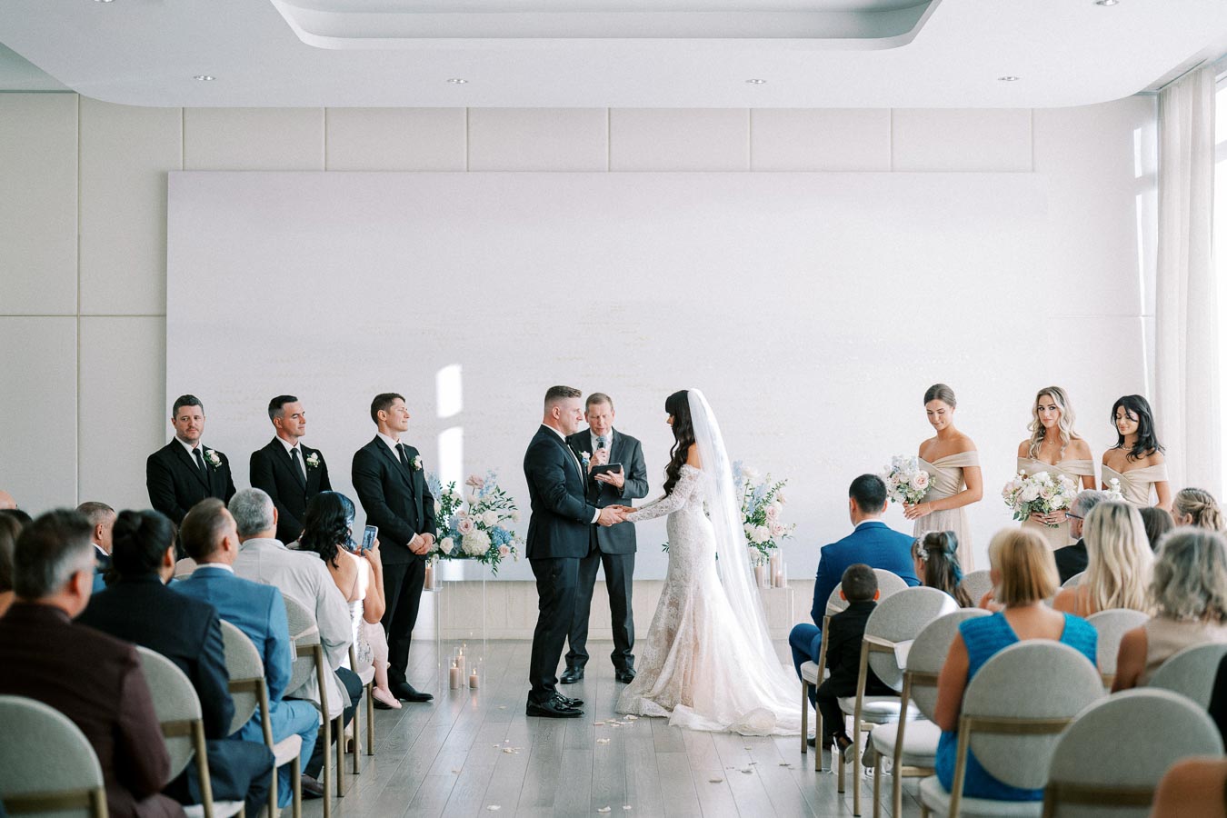 Elegant indoor wedding ceremony featuring a bride in a white gown and veil exchanging vows with the groom in a black suit; surrounded by bridesmaids in light beige dresses and groomsmen in formal attire, with seated guests observing.