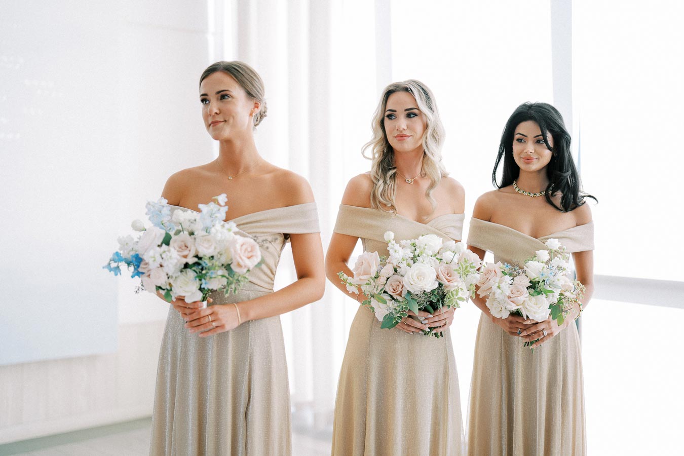 Three bridesmaids in elegant off-shoulder champagne dresses holding pastel floral bouquets, standing in a bright room.