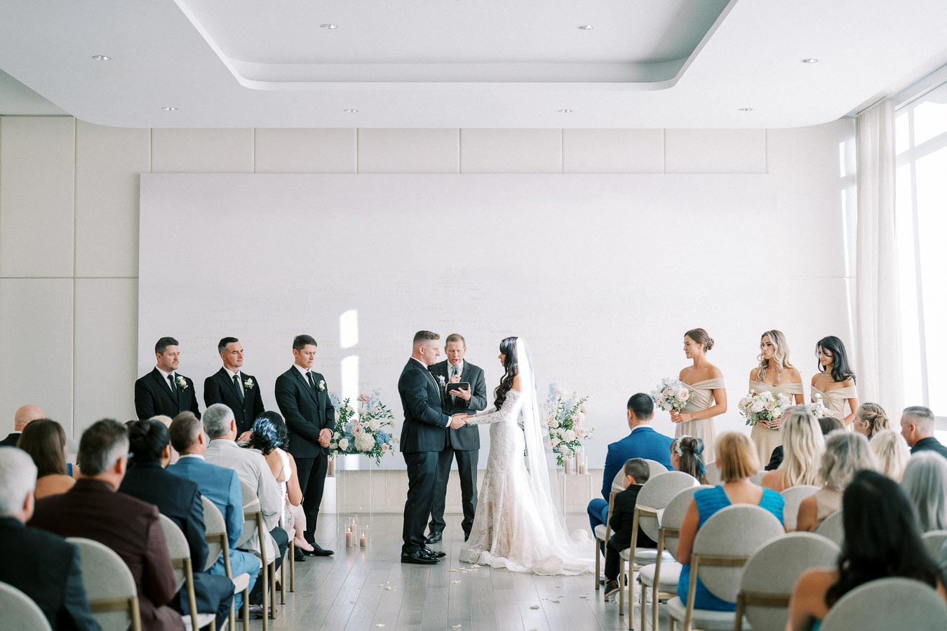 Elegant wedding ceremony in a bright, modern venue with the bride and groom exchanging vows. Bridesmaids in beige dresses and groomsmen in black suits stand by, while guests watch the intimate moment.