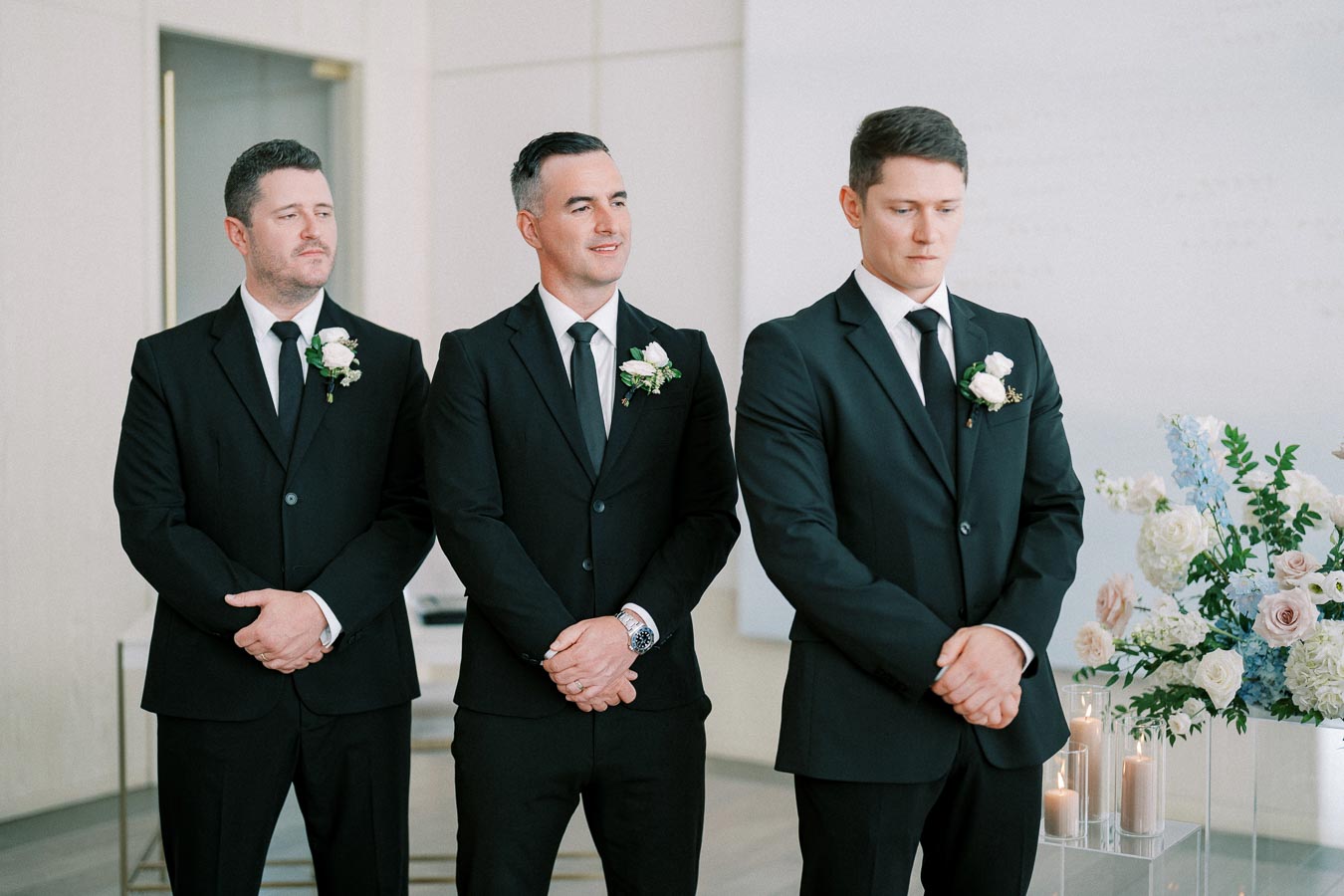 Three groomsmen in black suits and ties standing together indoors, with floral arrangements and candles in the background.
