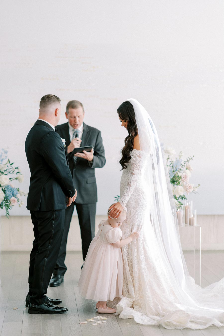 Bride and groom during wedding ceremony with a child hugging the bride's leg, elegant floral arrangements and candles in the background.