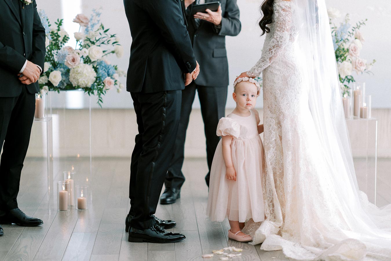 Elegant wedding scene with a bride in a lace gown gently placing her hand on a young flower girl's head, surrounded by groomsmen in black suits, soft floral arrangements, and candlelit decor.