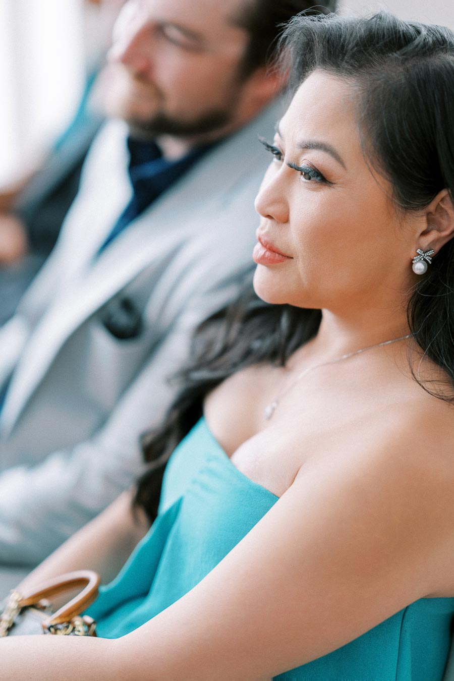 A woman in a turquoise dress attentively listening during an event, with a man in the background wearing a light gray suit.