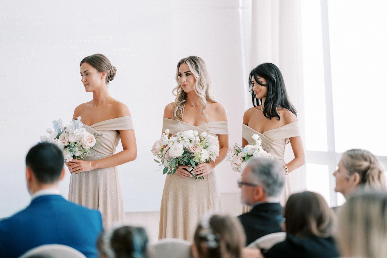 Three bridesmaids in elegant beige dresses stand holding bouquets of pastel flowers during a wedding ceremony, with seated guests in the foreground.