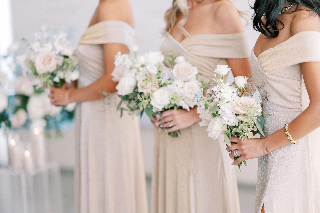 Three bridesmaids in elegant beige dresses holding pastel floral bouquets at a wedding ceremony.