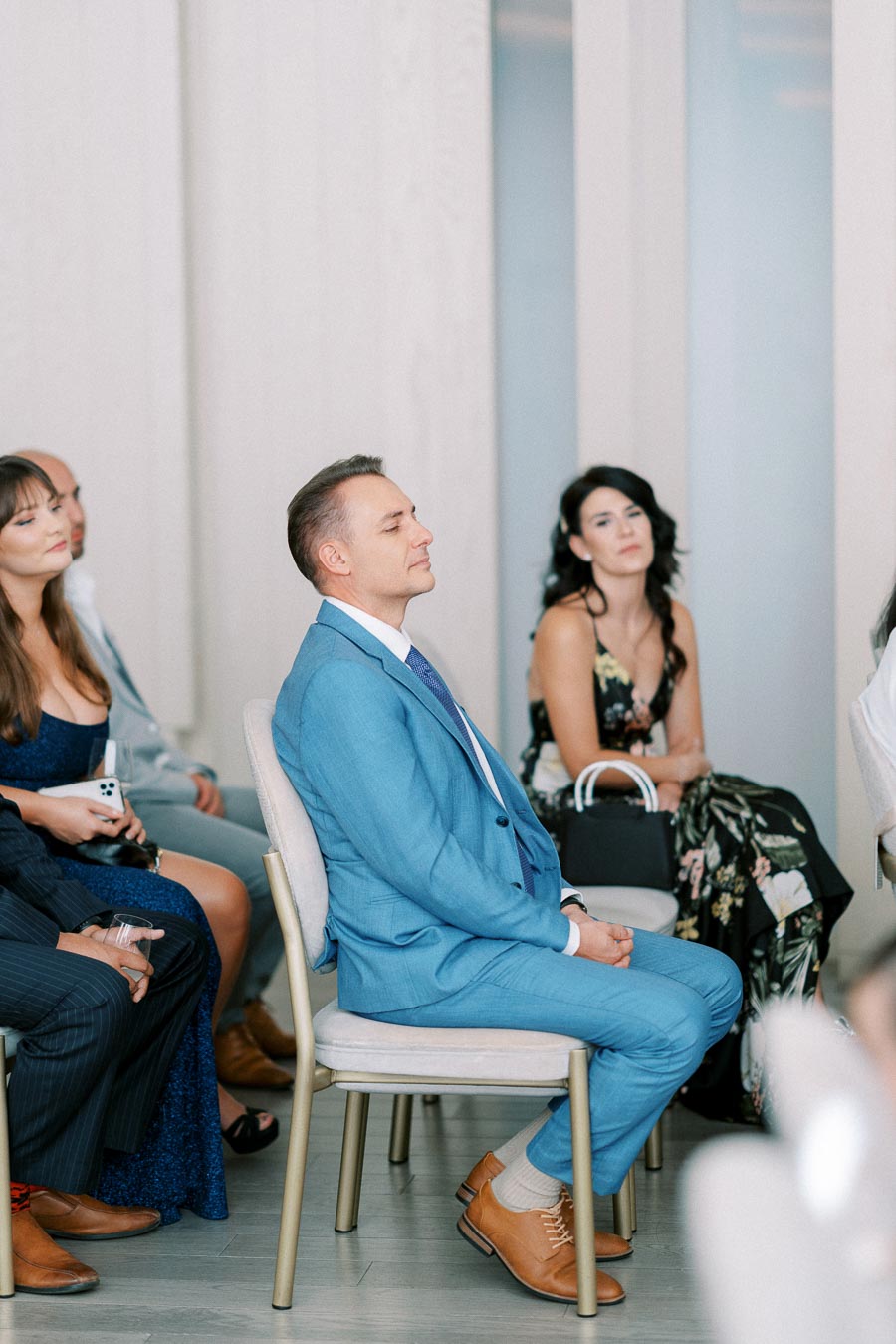 A group of people attentively listening to a presentation at an event, with a man in a blue suit sitting in the foreground.
