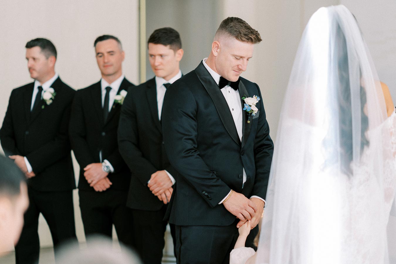 Groom in a black tuxedo standing with groomsmen during a wedding ceremony, holding hands with the bride, who is wearing a veil.
