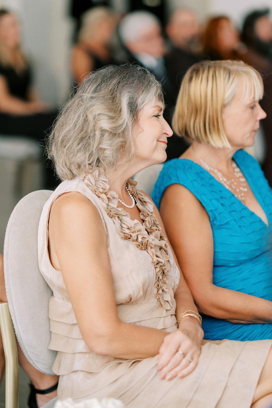 Women elegantly dressed sitting at a formal event, wearing beige and blue dresses with floral and textured details, showcasing stylish fashion and accessories.