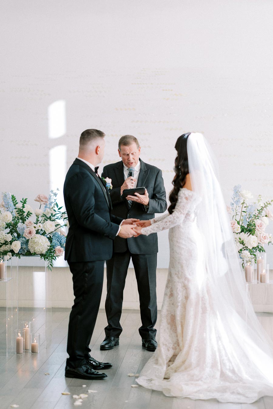 Wedding ceremony with bride and groom exchanging vows, wearing formal attire, in a venue adorned with elegant floral arrangements and candles, with an officiant reading from a book.