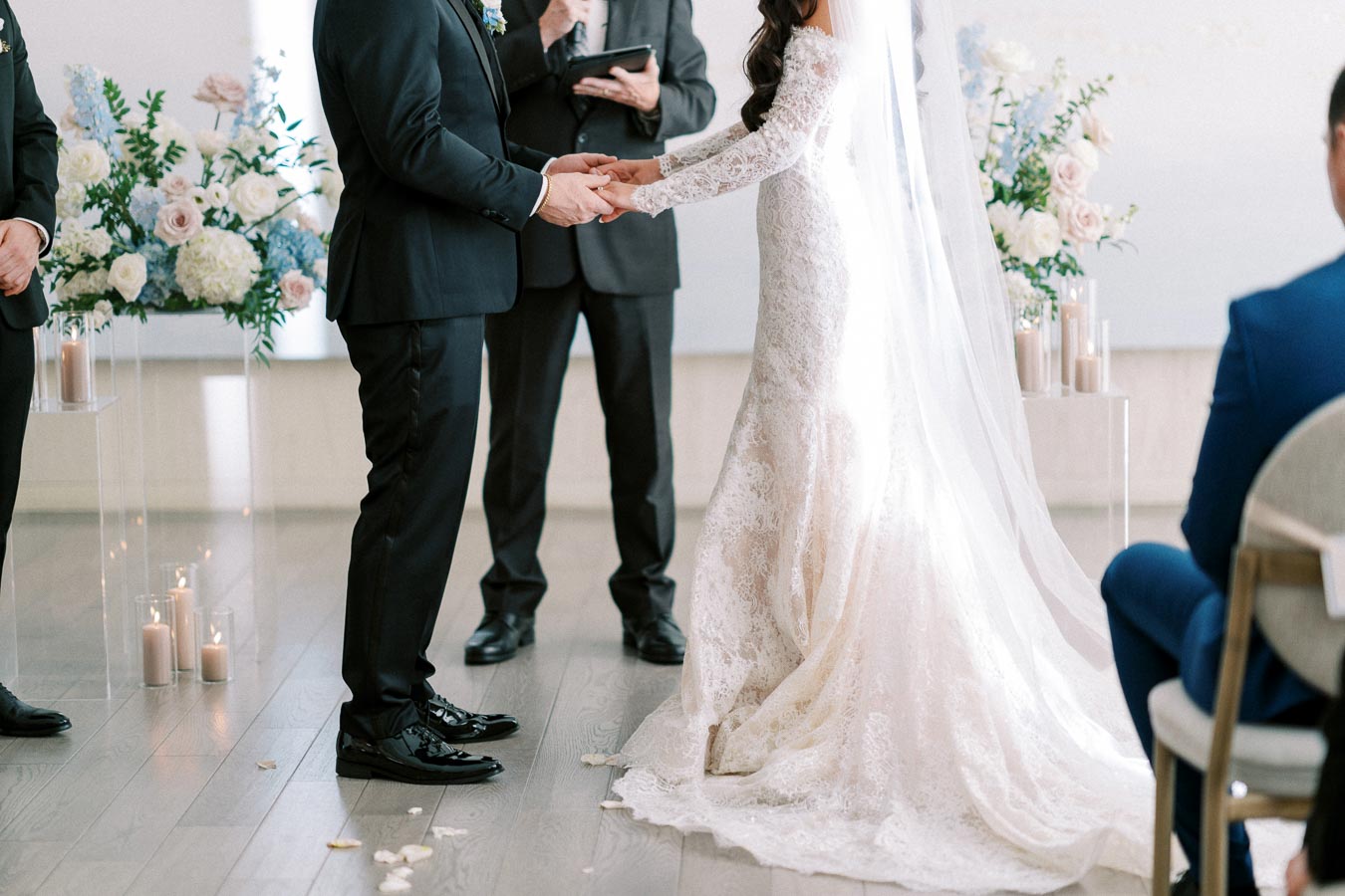 A bride and groom holding hands during a wedding ceremony, with the bride in an elegant lace gown and the groom in a black suit, surrounded by floral arrangements and candles.