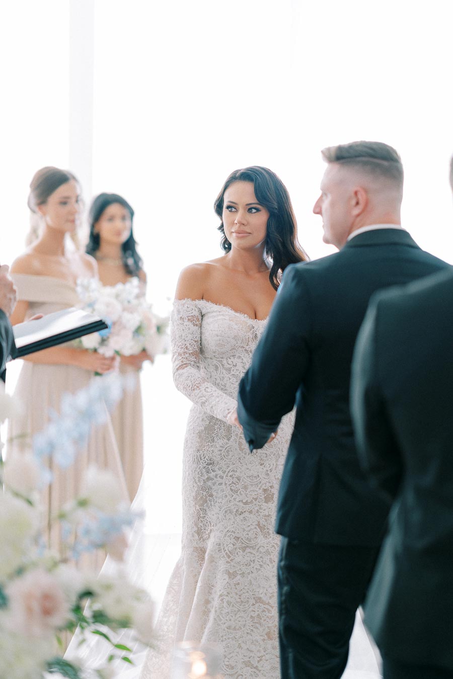 Wedding ceremony with bride in lace gown holding hands with groom, surrounded by bridal party in a floral-decorated venue.